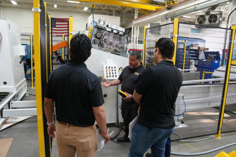 Three men, all dressed in black polo shirts, are gathered around an industrial machine inside a hop. One of them, a technician, is explaining the critical operation of the machine, holding a remote control with coiled wiring. The machine features a complex set of gears and a control panel with multiple buttons and switches. The surrounding area is clean and fenced off with yellow and black barriers for safety. In the background, an American flag hangs on the wall, and the factory is filled with other machinery and equipment. The point of the image is to display the professionalism of keeping a clean shop as critical for success.