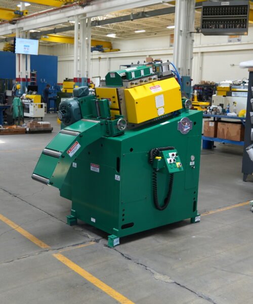 A large green and yellow industrial machine sits prominently in the center of a clean shop floor, with surrounding equipment and workbenches visible in the background. The factory has high ceilings, overhead cranes, and bright lighting. A worker in a yellow safety vest and overalls can be seen in the distance, inspecting another machine. The floor is marked with yellow lines for safety, and various tools and materials are organized on nearby shelves. The environment suggests heavy machinery production or maintenance work in a manufacturing facility.