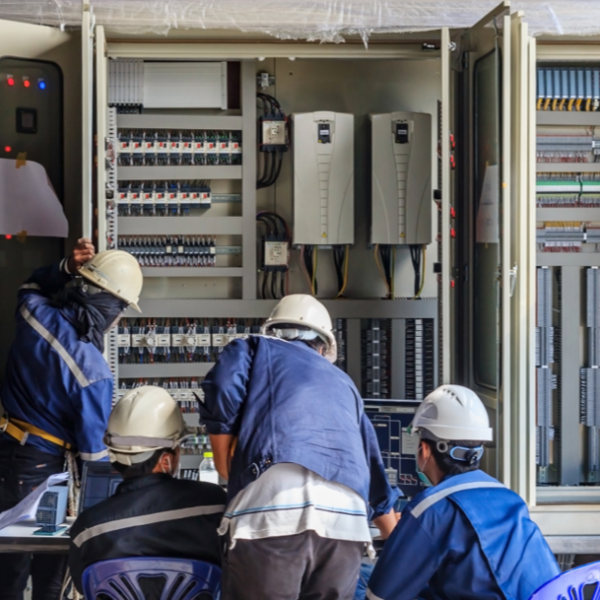 Technicians wearing safety helmets and work uniforms inspect and troubleshoot an industrial electrical control panel filled with wiring, circuit breakers, and programmable controllers.