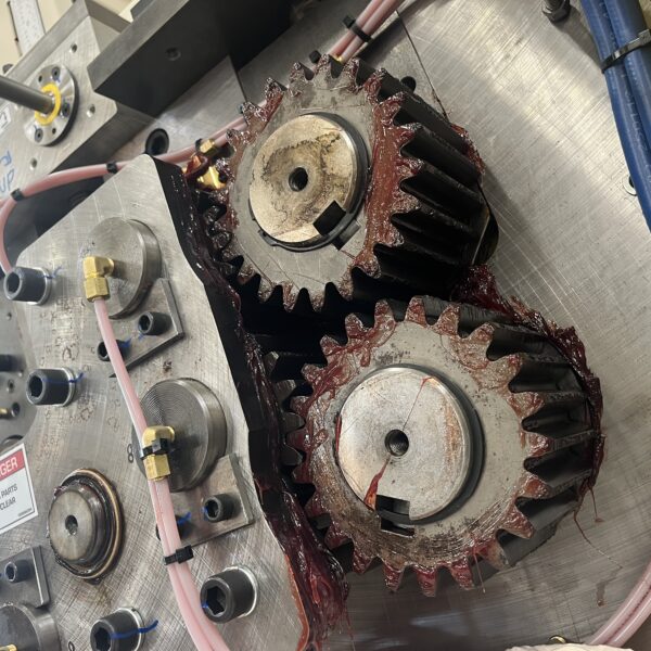 Close-up view of industrial machine gears coated in red grease, with hydraulic and pneumatic tubing connected to the surrounding metal assembly. Safety warning labels indicate moving parts.
